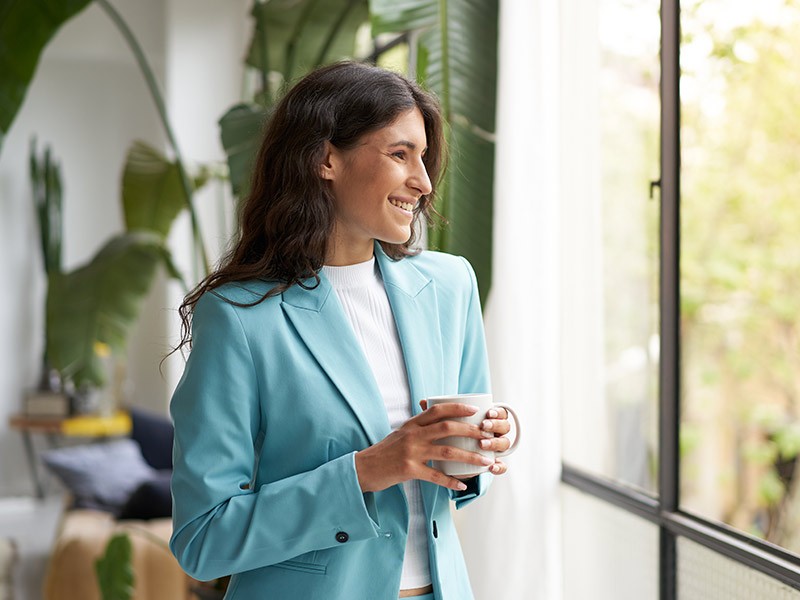 Young smiling business woman holding cup of coffee or tea and looking out the window