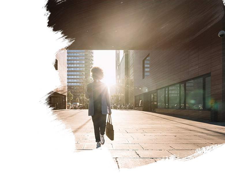 Woman walking on a city street between buildings with sun behind her