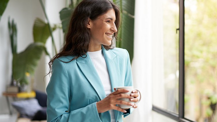 Young smiling business woman holding cup of coffee or tea and looking out the window