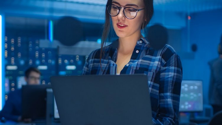 Portrait of a Smart Seductive Young Woman Wearing Glasses Holds Laptop. In the Background Technical Department Office with Specialists Working and Functional Data Server Racks