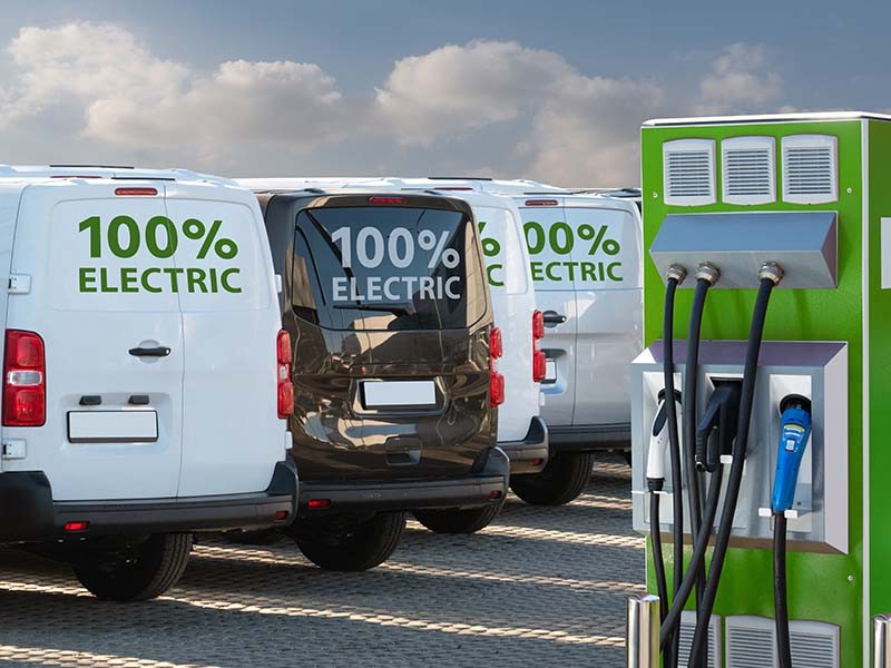 A fleet of vans that say “100% electric” are parked near an electric vehicle charging station