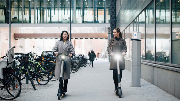 Two business women riding electric scooters on sidewalk in business district