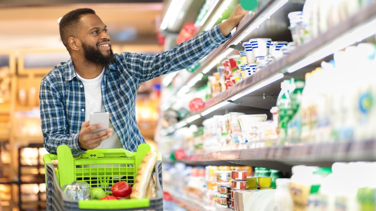 Male Buyer Shopping Groceries In Supermarket Taking Dairy Product From Shelf Standing With Shop Cart Indoors. Guy Buys Grocery Choosing Food In Super Market. Empty Space