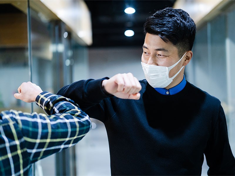 Two colleagues in face masks avoid handshake and greeting with bumping elbows at office workplace