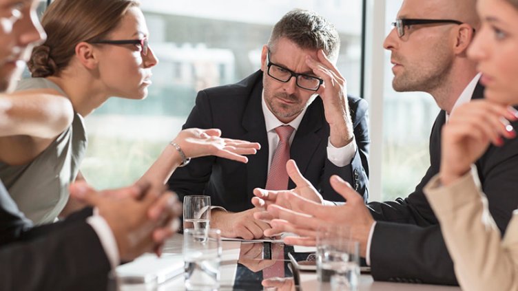Male executive sits at a table looking stressed as his business colleagues argue in a meeting around him